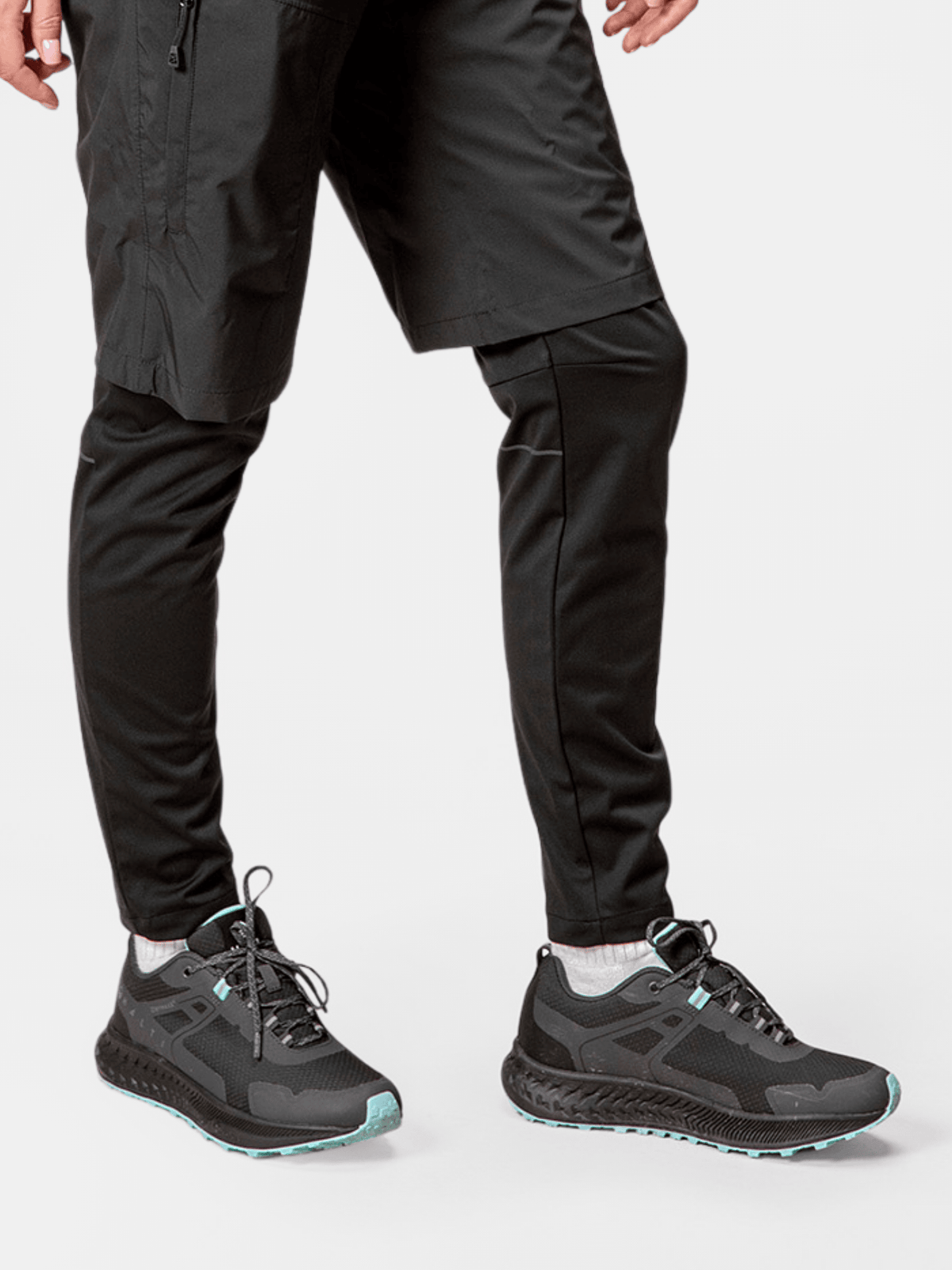 Henkilö, jolla on Halti Pallas Low 2 DX Hybrid Lenkkari Naisten kengät, mustat housut ja shortsit, seisoo valkoista taustaa vasten.