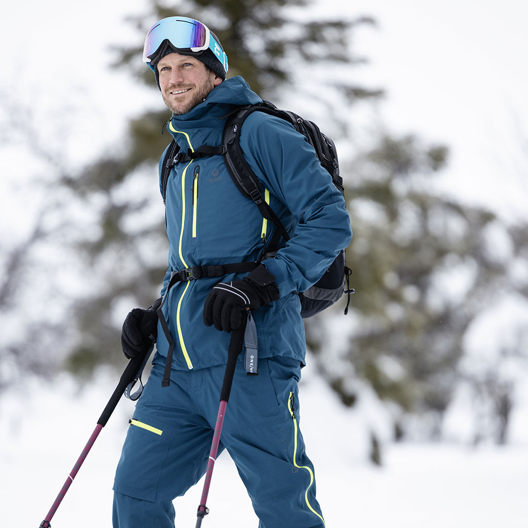 Sinisiin Halti Alpine 3L DX Kuorihousut Miesten ja suojalaseihin pukeutunut henkilö seisoo lumessa, pitää suksisauvoja ja hymyilee.