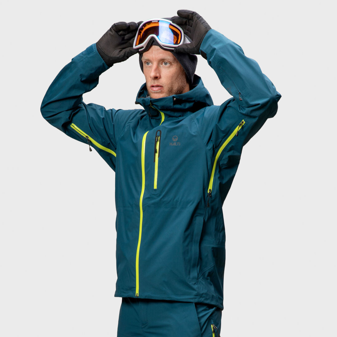 Henkilö, jolla on tiilenpunainen Halti Alpine 3L DX Kuoritakki Unisex ja hanskat, pitää hiihtolaseja päänsä yläpuolella vaaleaa taustaa vasten.