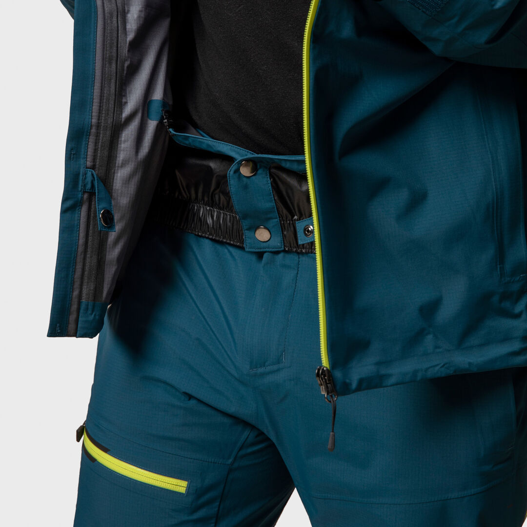 Henkilö, jolla on tiilenpunainen Halti Alpine 3L DX Kuoritakki Unisex ja keltaisilla vetoketjuilla varustetut ulkoiluhousut, kuvassa olkapäästä reiden puoliväliin.