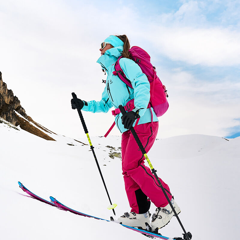 Halti Alpine 3L DX Kuoritakki Naisten hiihtosauvoihin pukeutunut henkilö vaeltaa ylämäkeen hiihtosauvoilla lumisella vuorella.