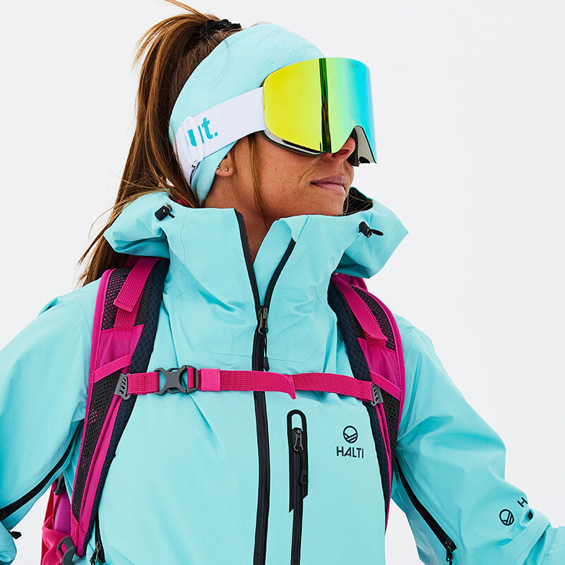 Henkilö vaaleansinisessä Halti Alpine 3L DX Kuoritakki Naisten ja suojalasit vaaleanpunainen reppu, kasvot sivuttain valkoinen.