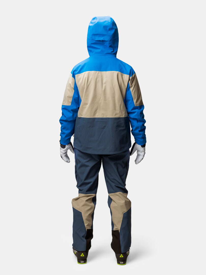 Takakuva henkilöstä, jolla on yllään sininen, beige ja tummanharmaa Halti Rizle Unisex 3L DX -laskettelutakki, housut, käsineet ja saappaat.