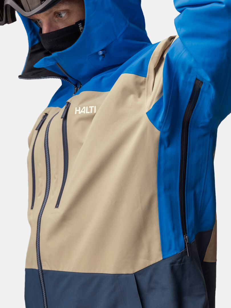 Henkilö, jolla on yllään sininen, beige ja tumman laivastonsininen Halti Rizle Unisex 3L DX -laskettelutakki, jossa on vetoketjullinen tuuletusaukko kainalossa.