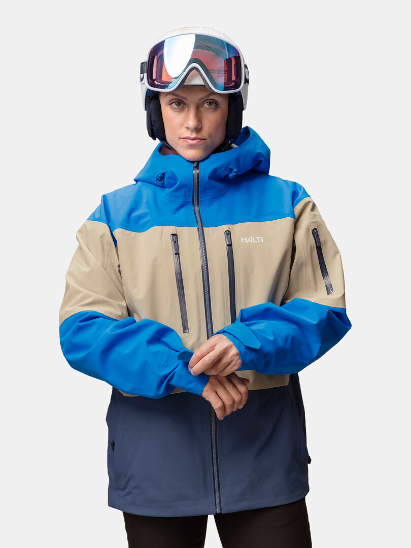 Henkilö, jolla on päällään hiihtokypärä, suojalasit sekä sininen ja beige Halti Rizle Unisex 3L DX -laskettelutakki.