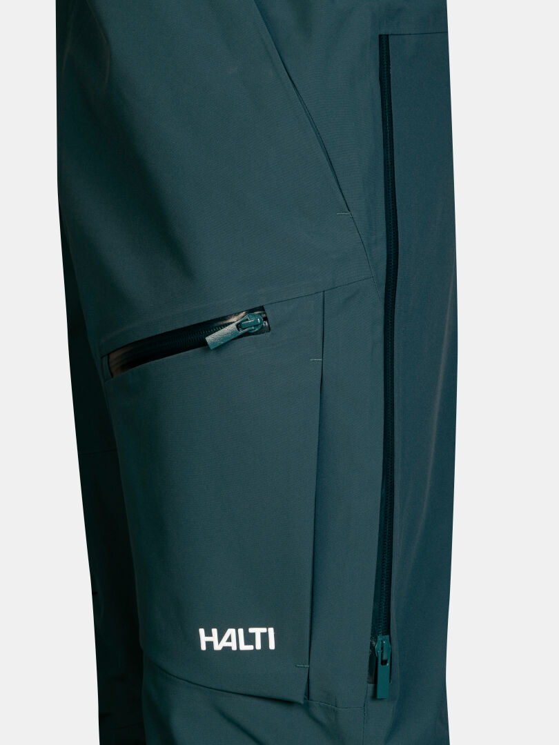 Tumman sinivihreät Halti Rizle 3L DX -lasketteluhousut (unisex), joissa on vetoketjullinen reisitasku ja sivutuuletusaukot.