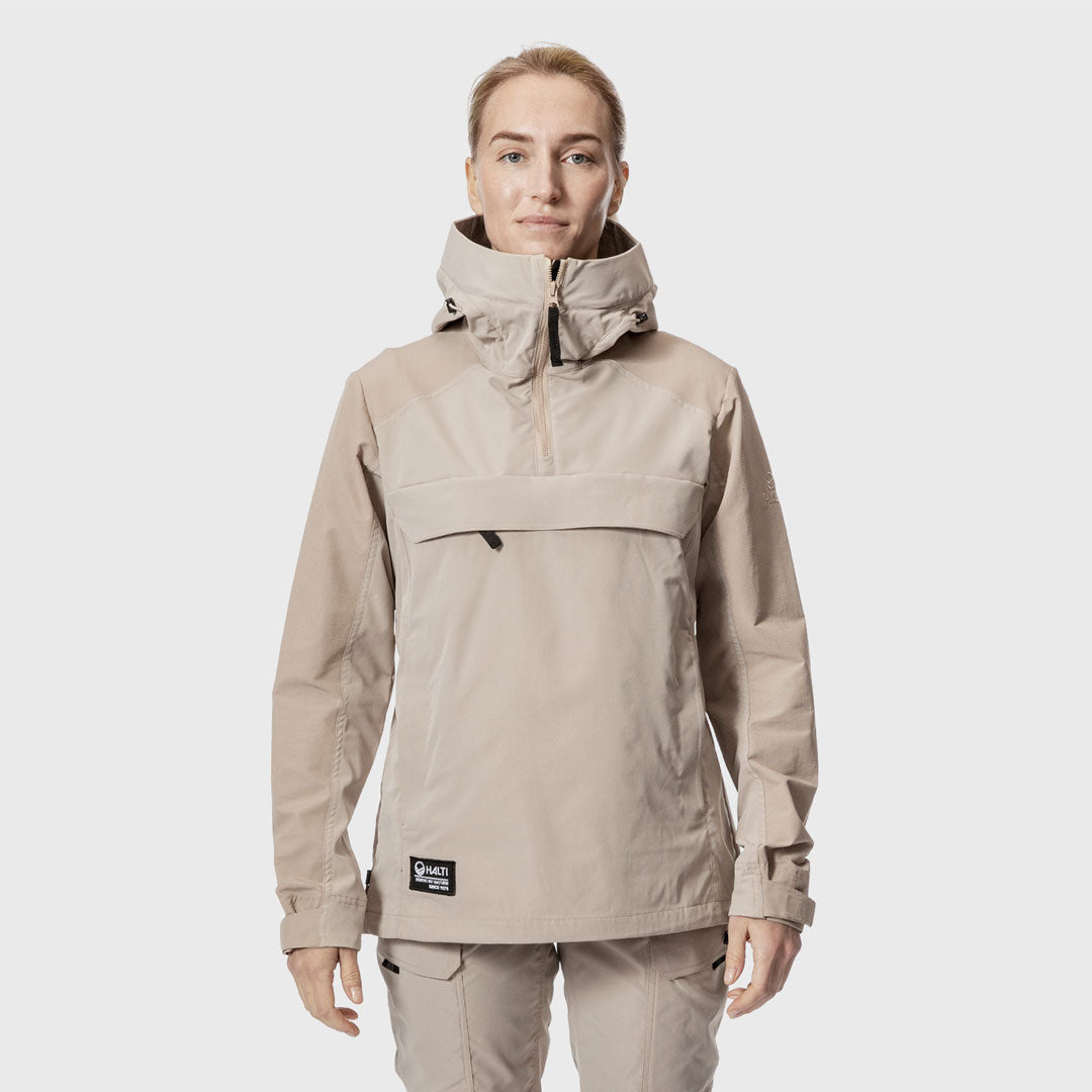 Henkilö, jolla on beige hupullinen takki ja siihen sopivat housut, seisoo kasvot menosuuntaan valkoista taustaa vasten.