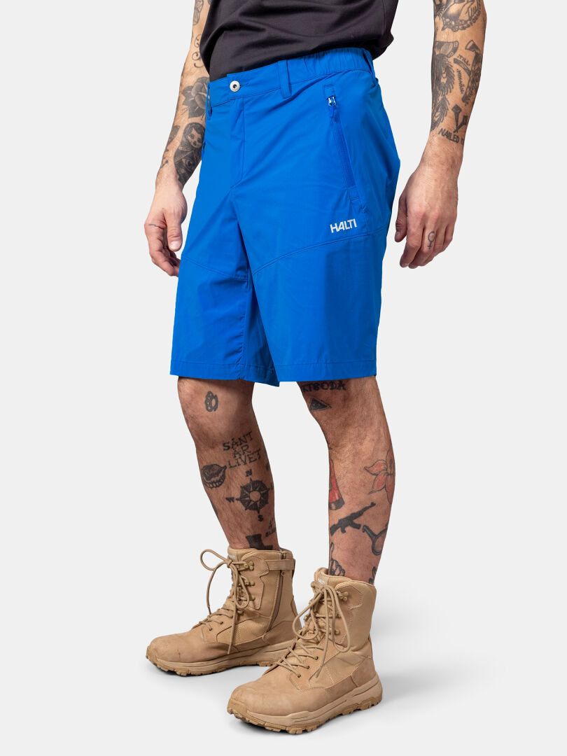 Henkilöllä on kirkkaan sininen Halti Pallas Cool X-stretch Shortsit Miesten, musta paita, ruskehtavat saappaat ja tatuoinnit.