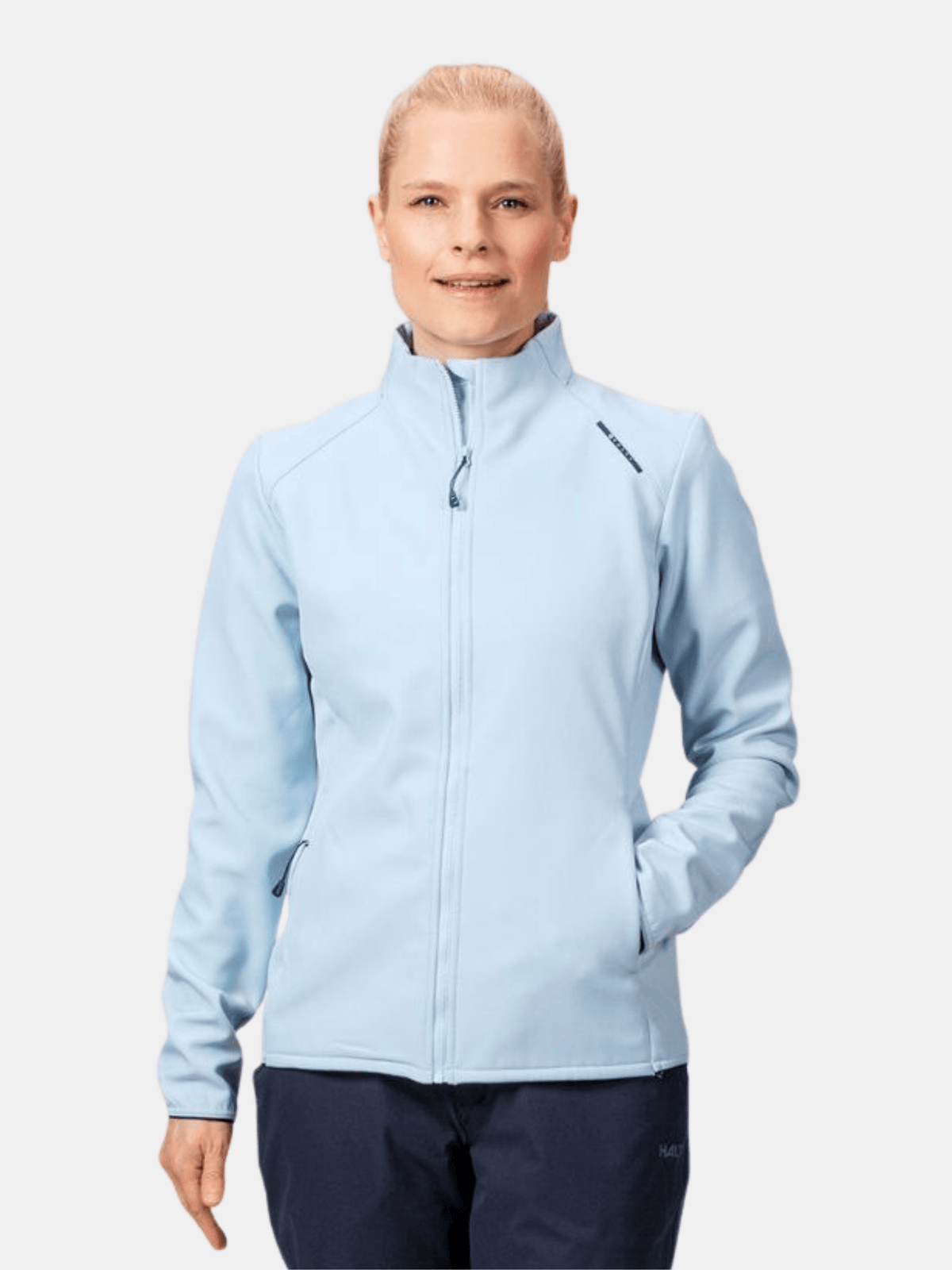 Nainen seisoo yksinkertaista valkoista taustaa vasten, kädet taskuissa, yllään Halti Fore Naisten Softshell Takki.