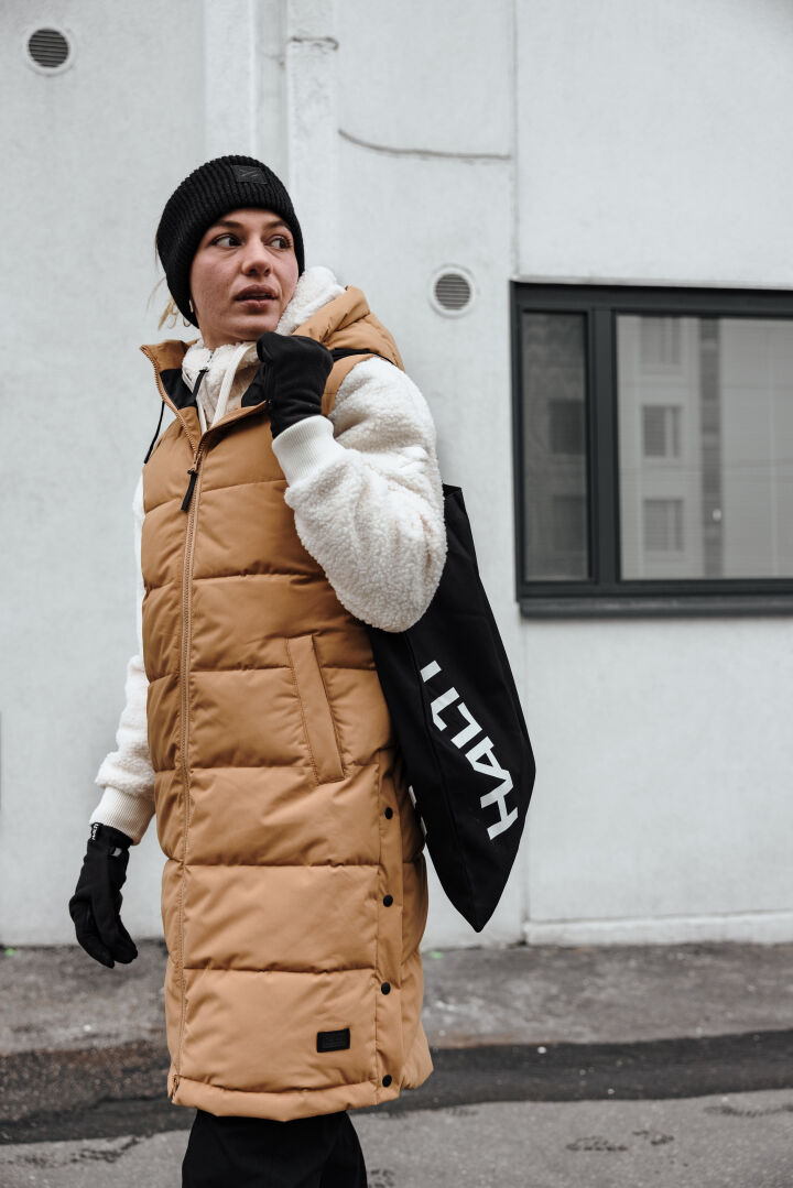Henkilö, jolla on ruskeanruskea puskuripuku, Halti Lykky Fleecepusero Naisten ja musta hattu, kantaa ulkona mustaa, valkoisella tekstillä varustettua kasettia.