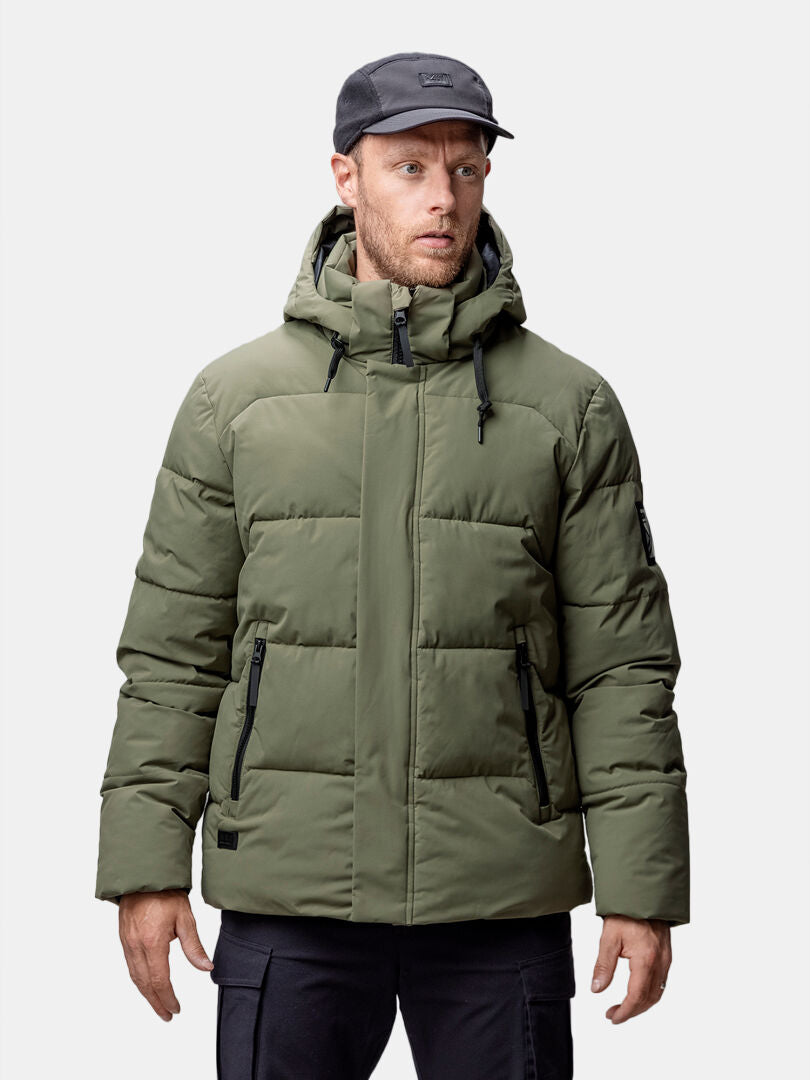 Halti Brahe Puffer Talvitakki, musta lippis ja housut päällä oleva mies seisoo vaaleaa taustaa vasten.