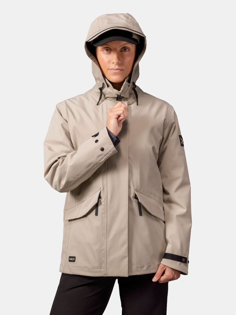 Henkilö, jolla on beige Halti Tokoi DX Takki Naisten ja mustat housut, seisoo edessä ja pitää toisella kädellä kiinni takin vetoketjusta.