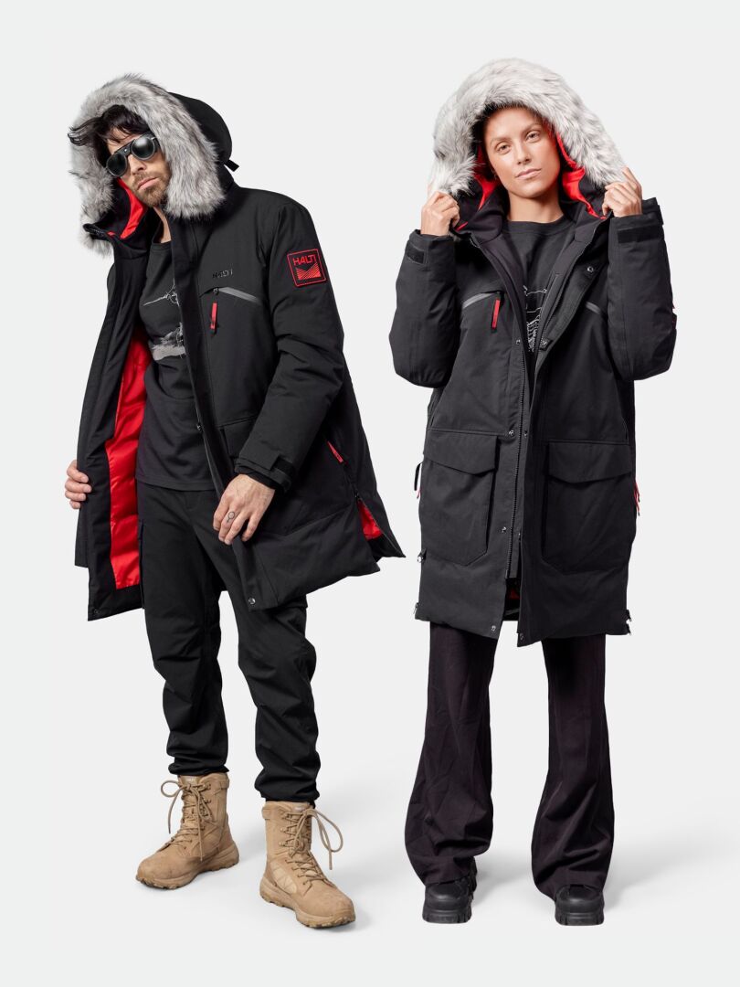 Kaksi ihmistä, joilla on Halti 1976 Down Parka Unisex ja mustat housut, seisovat valkoista taustaa vasten.