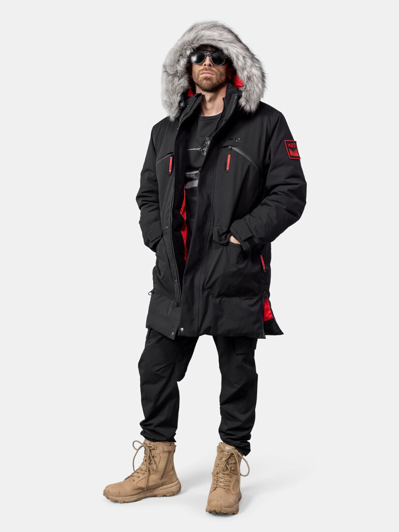 Henkilö, jolla on Halti 1976 Down Parka Unisex, aurinkolasit, mustat housut ja ruskehtavat saappaat, seisoo tavallista taustaa vasten.