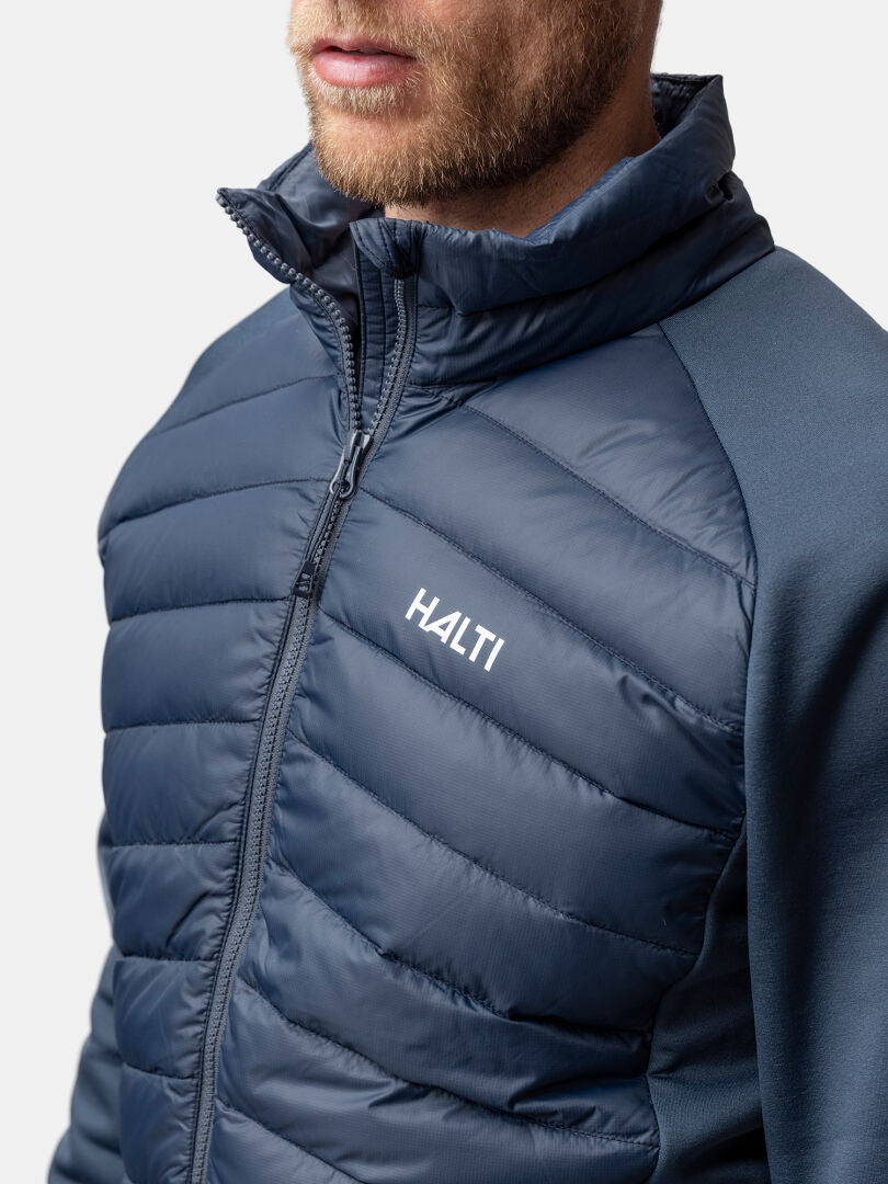 Mies, jolla on sininen Halti Huippu Hybrid Untuvatakki Miesten takki, kuvattuna olkapäistä ylöspäin tavallista taustaa vasten.