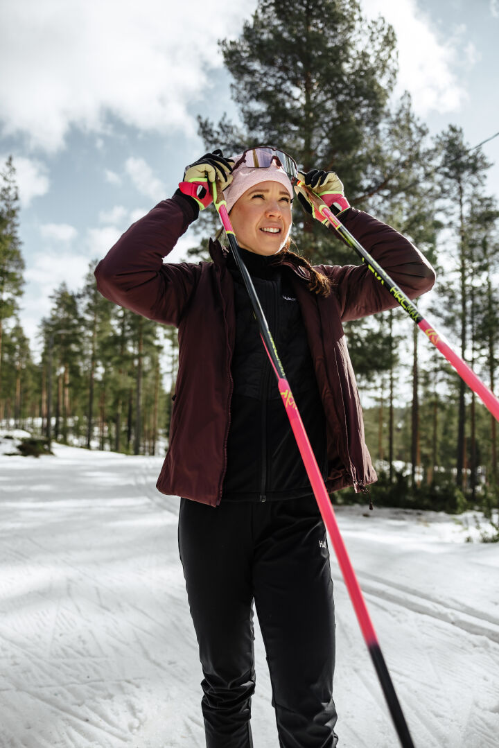 Halti Framover Hybrid Hiihtotakki Naisten henkilö seisoo lumisella ladulla hiihtosauvojen kanssa puiden välissä.