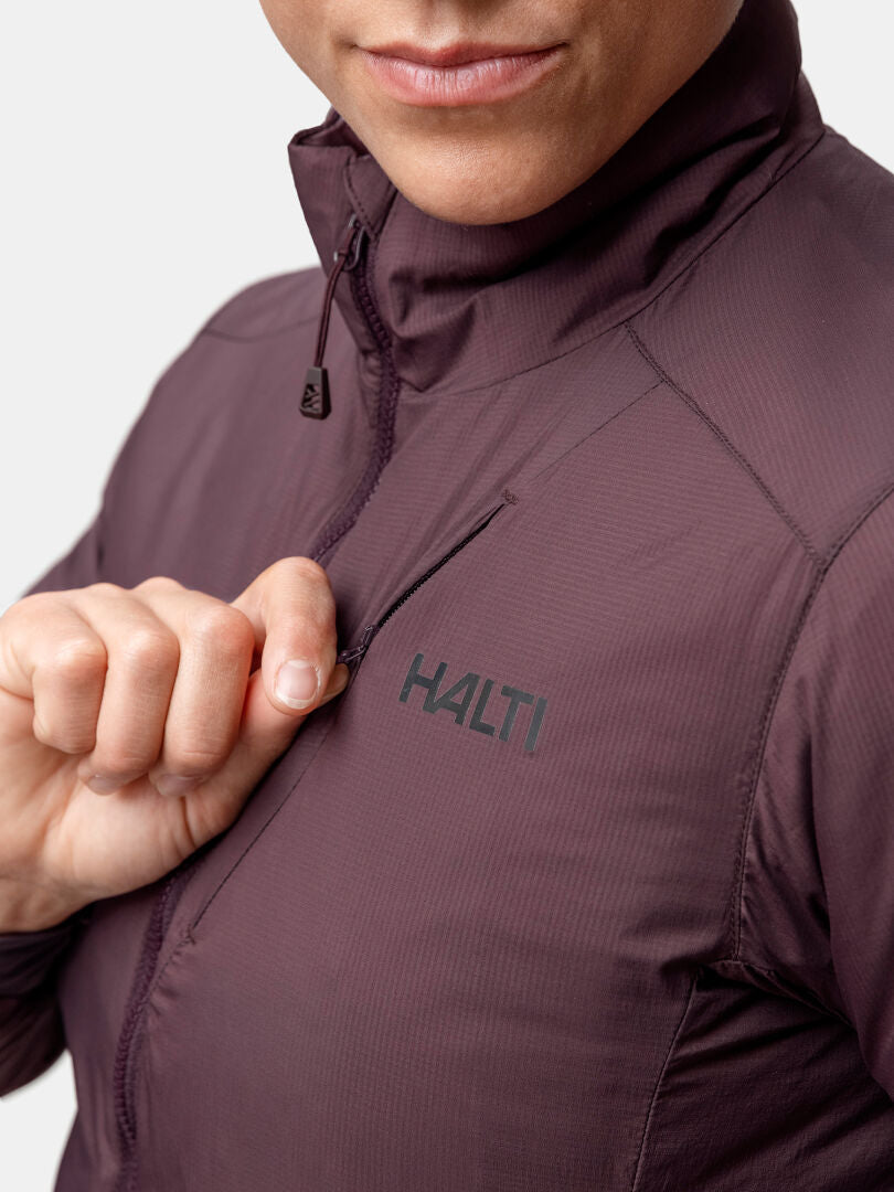 Henkilö vetää vetoketjun kiinni violetista Halti Haave Primaloft Takki Naisten, jonka logo näkyy rinnassa yksiväristä taustaa vasten.