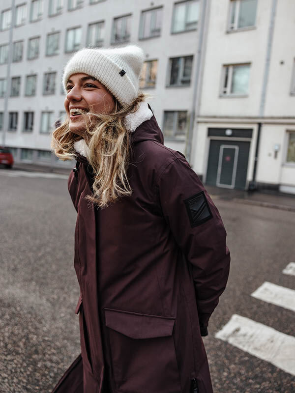 Henkilö, jolla on ruskeankeltainen takki ja Halti Vuolle Mohairpipo, hymyilee kaupungin kadulla, jonka taustalla on rakennuksia.