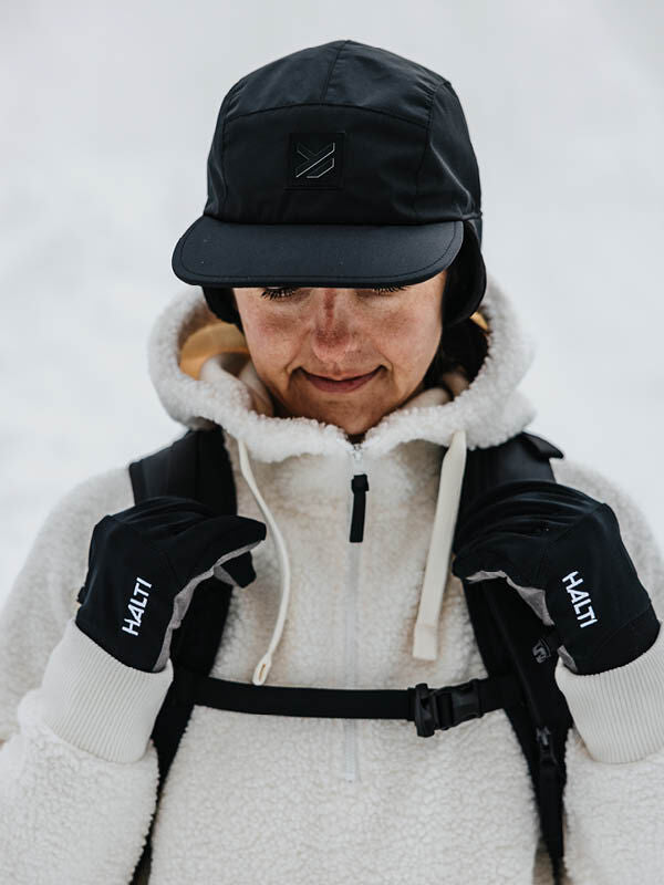 Henkilö, jolla on yllään musta Halti Skutta Lippis, valkoinen fleecetakki ja Halti-hanskat, seisoo ulkona lumessa.