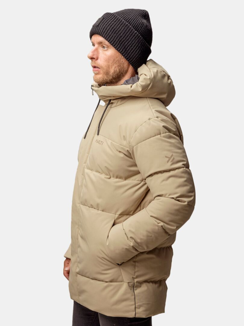 Mies, jolla on beige puserotakki ja Halti Huurre Pipo, seisoo vasemmalle päin vaaleaa taustaa vasten.