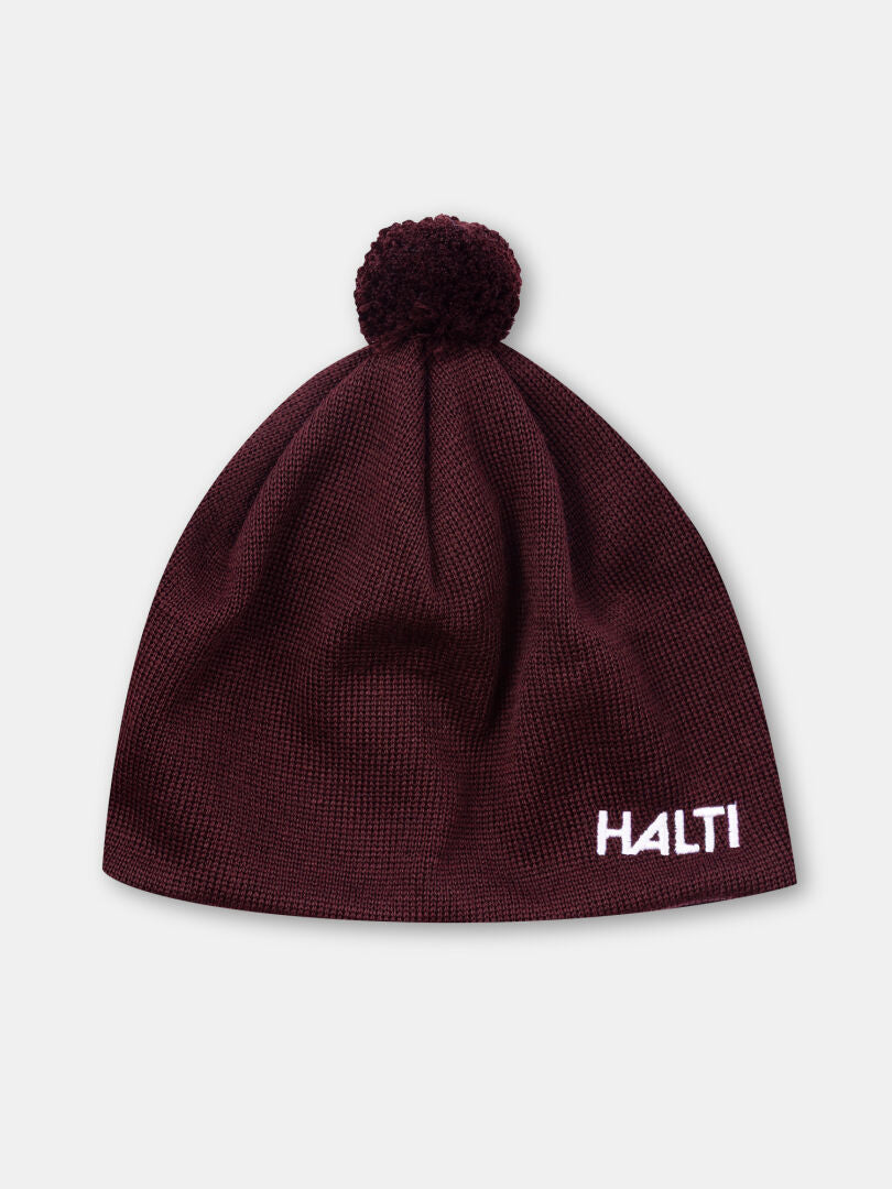 HALTIn ruskeankeltainen Peesi II Pipo merinovilla pipo, jossa on pompsupää ja valkoinen HALTI-kirjonta reunan lähellä.