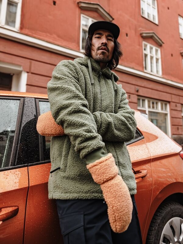 Halti Lykky Fleecehanskat ja suuret beiget hanskat yllään oleva mies seisoo oranssin auton vieressä punaisen rakennuksen edessä.