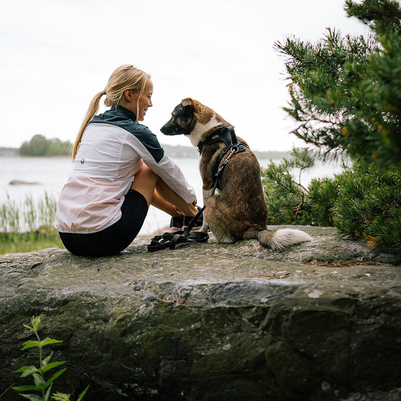 Halti Urbanite Naisten Treenitakki -takissa oleva nainen istuu ulkona kivellä kasvot koiraa kohti, takanaan puita ja vettä.