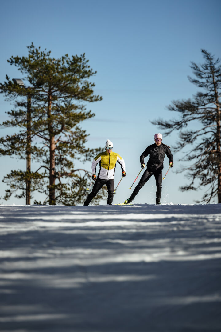 Kaksi ihmistä Halti Framover Hiihtotakki Miesten hiihtää lumisella ladulla kirkkaan sinisen taivaan alla.