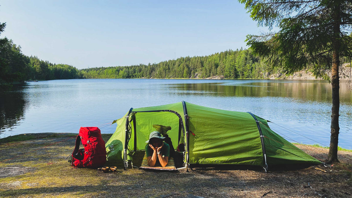Henkilö makaa vihreässä teltassa järven rannalla, punainen reppu lähellä, puiden ja tyynen veden ympäröimänä.