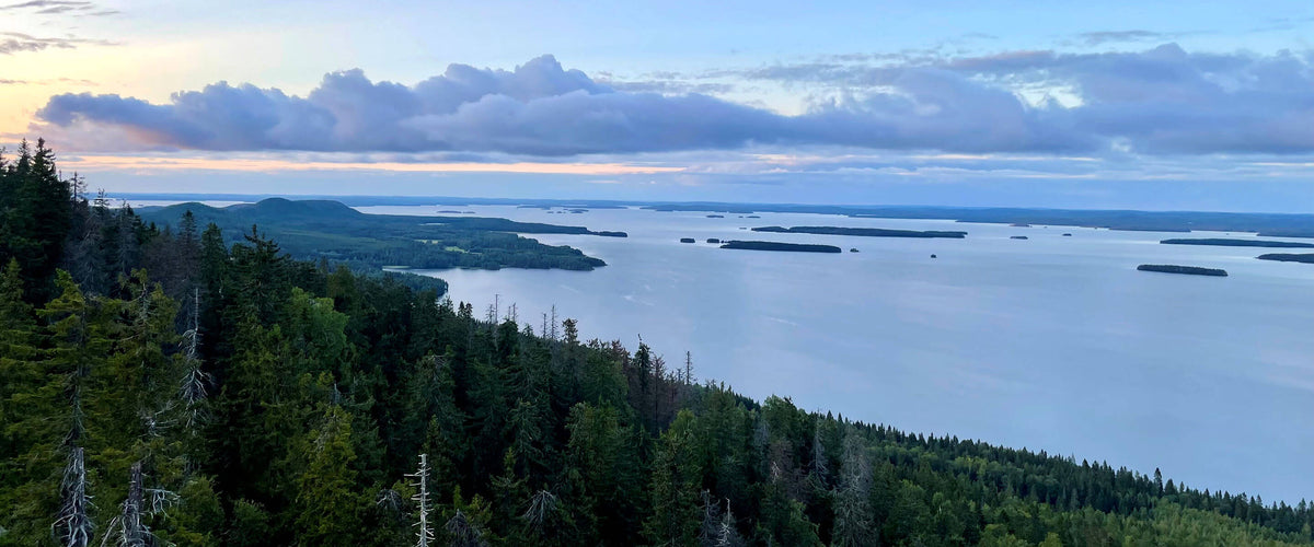 Näkymä suuresta järvestä, jossa on hajallaan olevia saaria, tiheän vihreän metsän ja pilvisen taivaan ympäröimänä auringonlaskun aikaan.