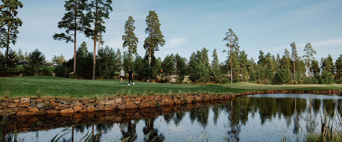 Kaksi ihmistä kävelee golfkentällä lammen lähellä, puiden ja kivimuurin ympäröimänä sinisen taivaan alla.