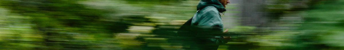 Vihreään takkiin pukeutunut henkilö juoksee hämärtyneen, vihreän metsän läpi, kuvattu motion blur -efektillä.