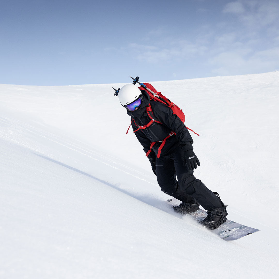 Henkilö, jolla on musta Halti Alpine 3L DX Kuorihousut Miesten ja punainen reppu, lumilautailee lumista rinnettä alas kirkkaalla taivaalla.