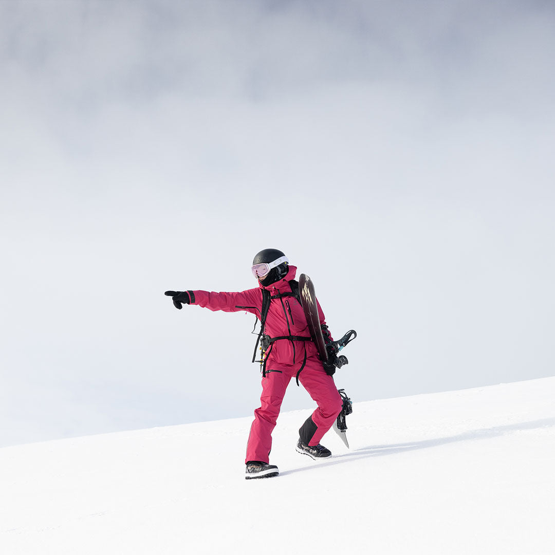 Henkilö punaisessa Halti Alpine 3L DX Kuoritakki Naisten kävelee lumella lumilauta kädessään ja osoittaa pilvisen taivaan alla.