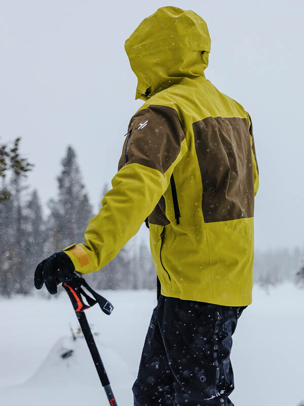 Henkilö, jolla on yllään limenvihreä ja ruskea Halti Rizle Unisex 3L DX Laskettelutakki, pitelee hiihtosauvoja lumisessa maisemassa.