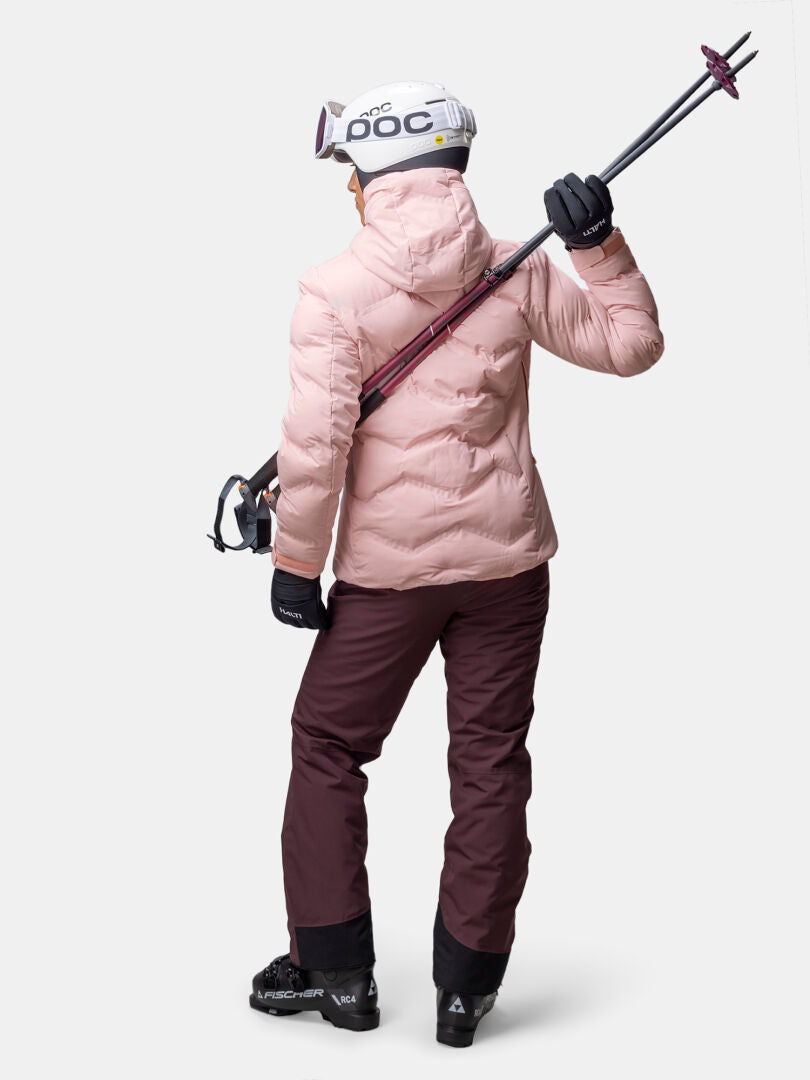 Henkilö, jolla on Halti Nordic Arcty II Laskettelutakki, ruskeat housut ja kypärä, pitää hiihtosauvoja olkapäänsä yli, kasvot poispäin.