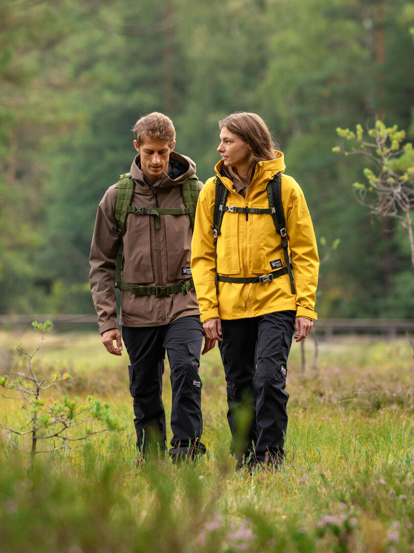 Kaksi Halti Hiker II DrymaxX Kuorihousut Naiset -takeissa ja repuissa olevaa ihmistä kävelee rinnakkain ruohoisella, puiden reunustamalla maastolla.