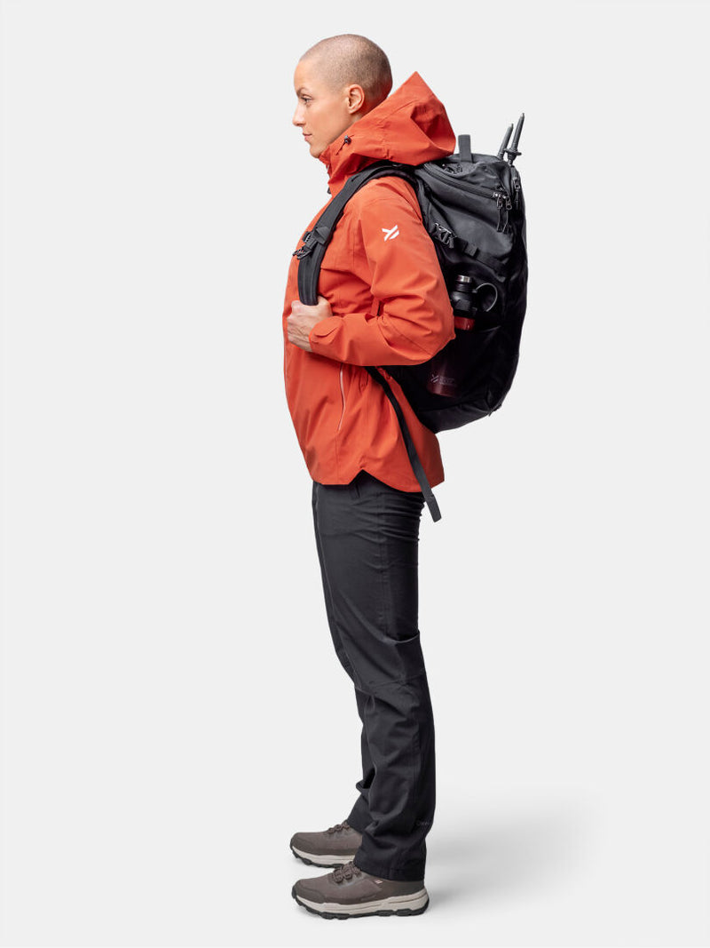 Henkilö, jolla on oranssi Halti Staala DX Kuoritakki Naisten, mustat housut ja vaelluskengät, seisoo profiilissa iso musta reppu mukanaan.