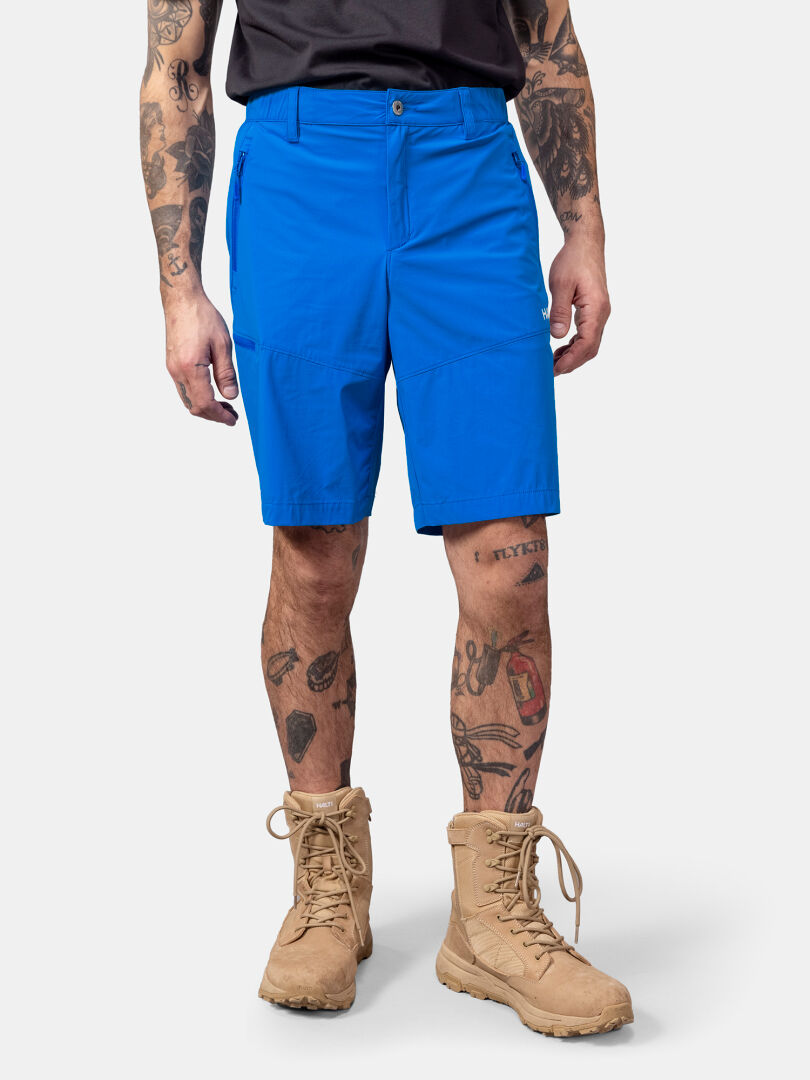 Miehellä Halti Pallas Cool X-stretch miesten shortsit siniset, ruskehtavat taistelusaappaat, musta paita; tatuoidut kädet ja jalat.