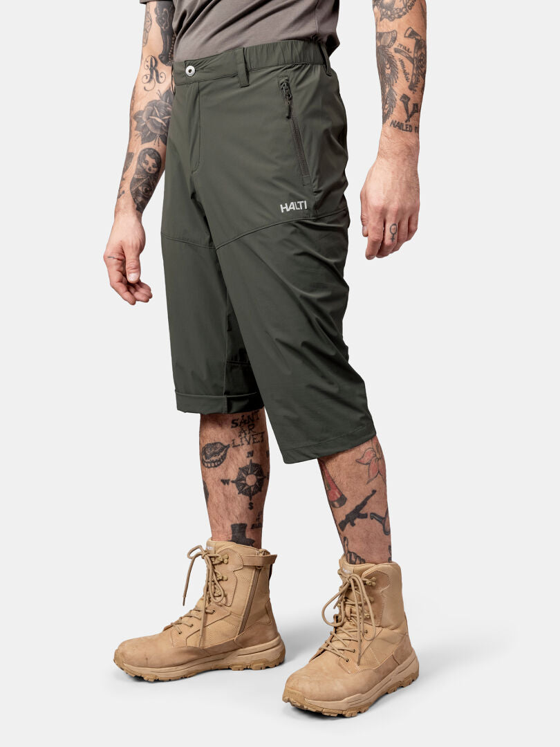 Tatuoidulla henkilöllä on oliivinvihreät Halti Pallas Cool X-stretch Caprit Miesten ja ruskehtavat saappaat valkoista taustaa vasten.