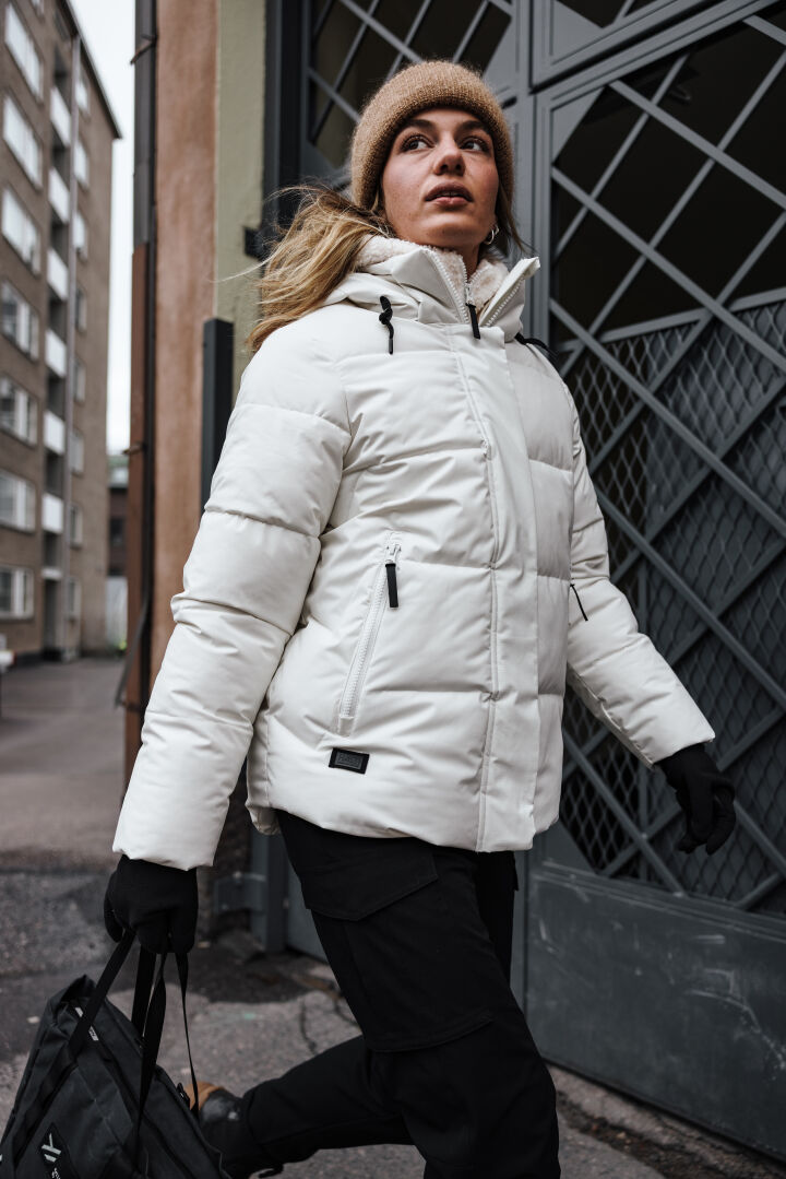 Joku Halti Brahe Puffer Talvitakki Naisten, mustat hanskat ja beige pipo kävelee ulos mustaa laukkua kantaen.