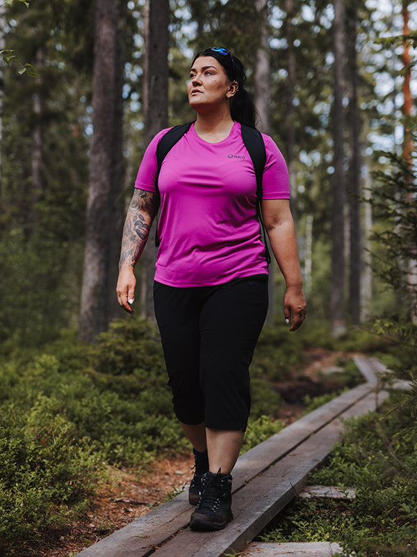Henkilö Halti Drive X-stretch Caprit Naisten Plus -mustissa housuissa kävelee metsäpolkua yllään vaaleanpunainen paita ja reppu.