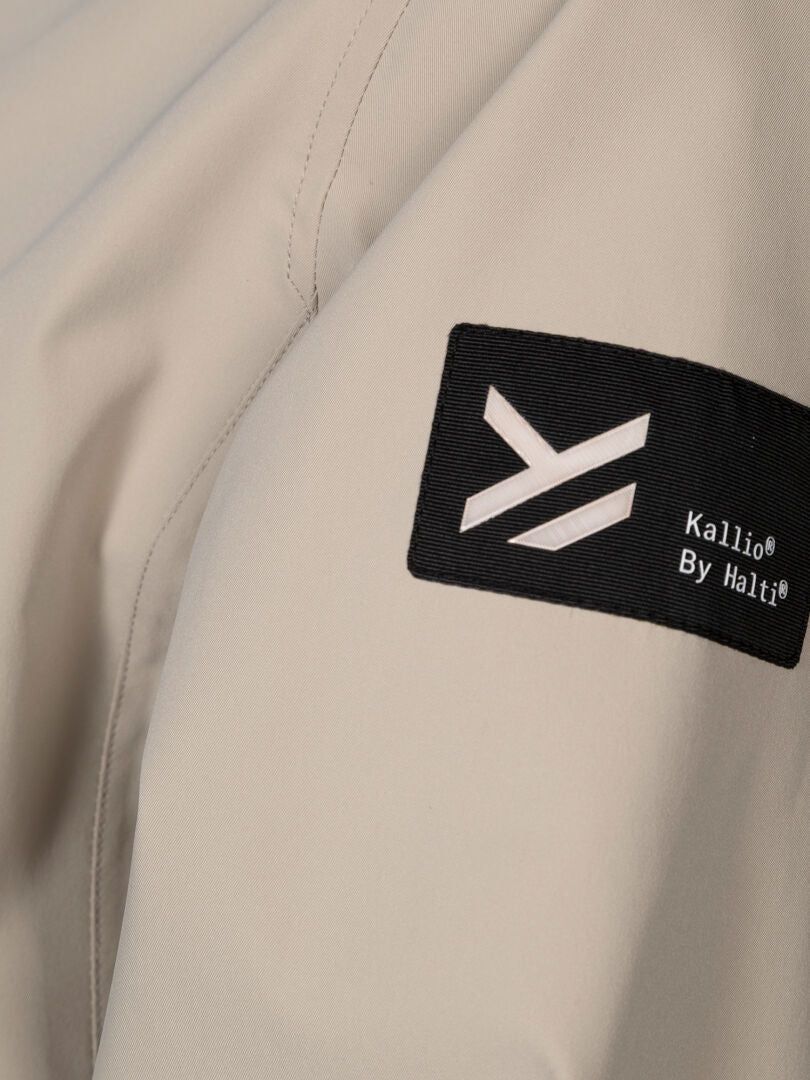 Lähikuva beigestä Tokoi DX Takki Naisten by Halti -paidasta, jossa on musta laastari ja valkoinen geometrinen logo.