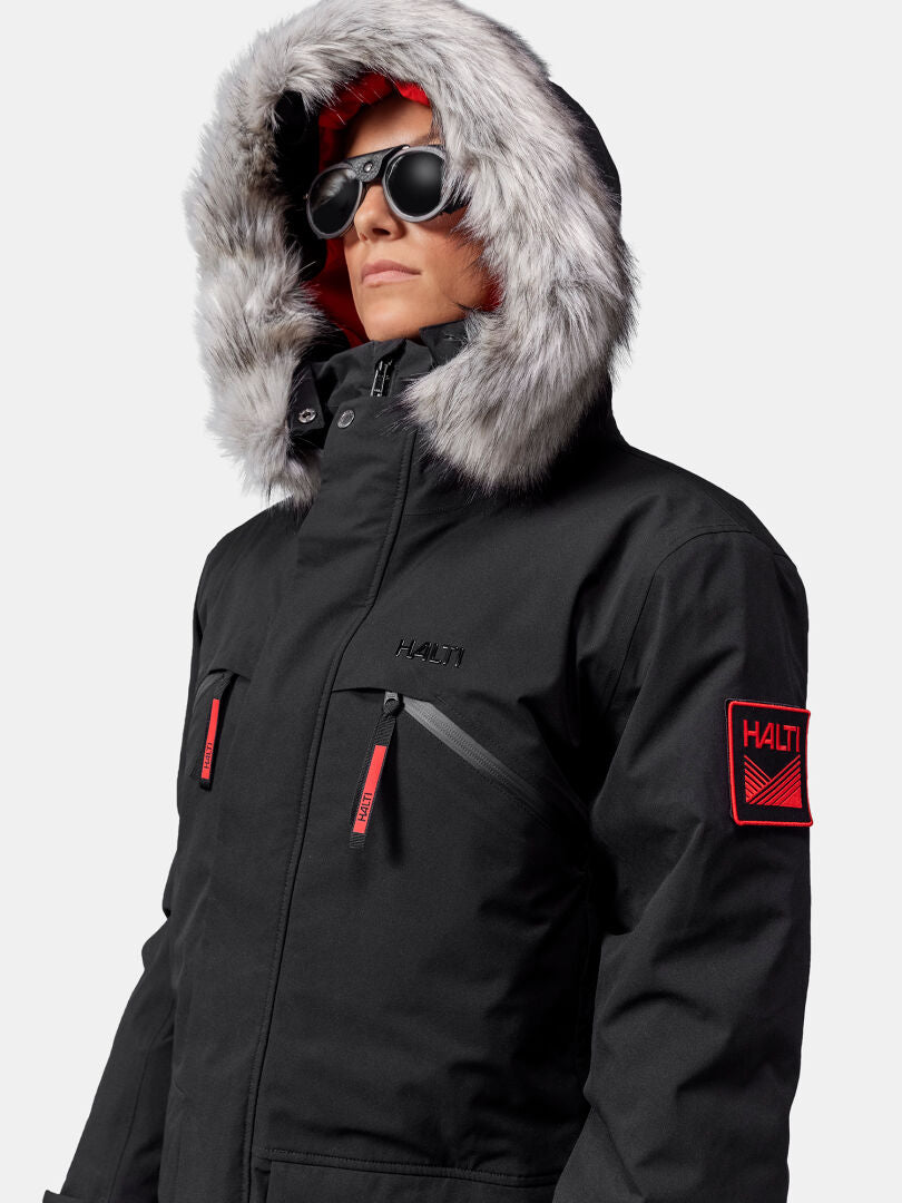 Päällään Halti 1976 Down Parka Unisex, jossa on turkisvuorillinen huppu, aurinkolasit ja Halti-laastari tavallista taustaa vasten.