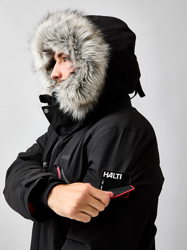 Henkilö lämmin talvitakki pitää HALTI-laastaria mustan Halti 1976 Down Parka Unisex -turkinsa hihassa.