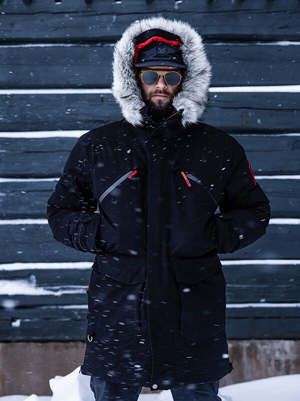 Henkilö, jolla on Halti 1976 Down Parka Unisex, seisoo laskevassa lumessa tumman puuseinän vieressä aurinkolasit ja hanskat yllään.