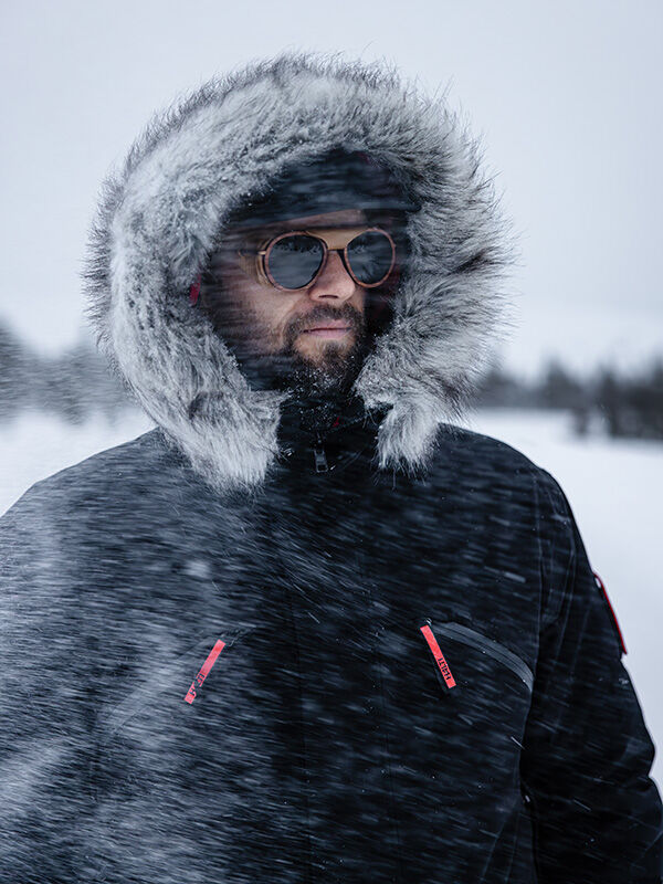 Henkilö, jolla on Halti 1976 Down Parka Unisex ja aurinkolasit, seisoo lumessa, kun tuuli puhaltaa lunta hänen kasvoilleen.