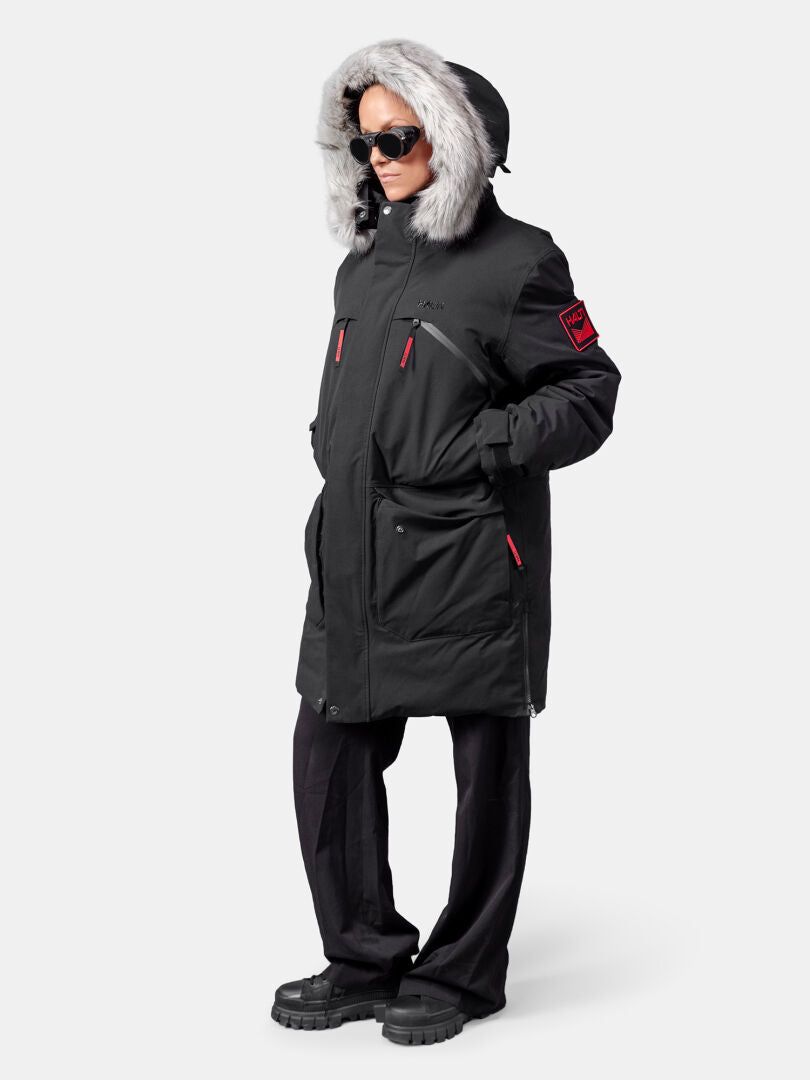 Henkilö, jolla on Halti 1976 Down Parka Unisex, mustat housut, tummat saappaat ja aurinkolasit tavallista taustaa vasten.