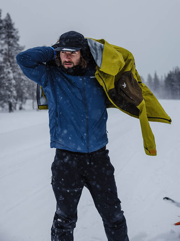 Talvivaatteisiin pukeutunut mies pukee keltaisen Halti Haave Primaloft Takki Miesten päälleen, kun ulkona sataa lunta.
