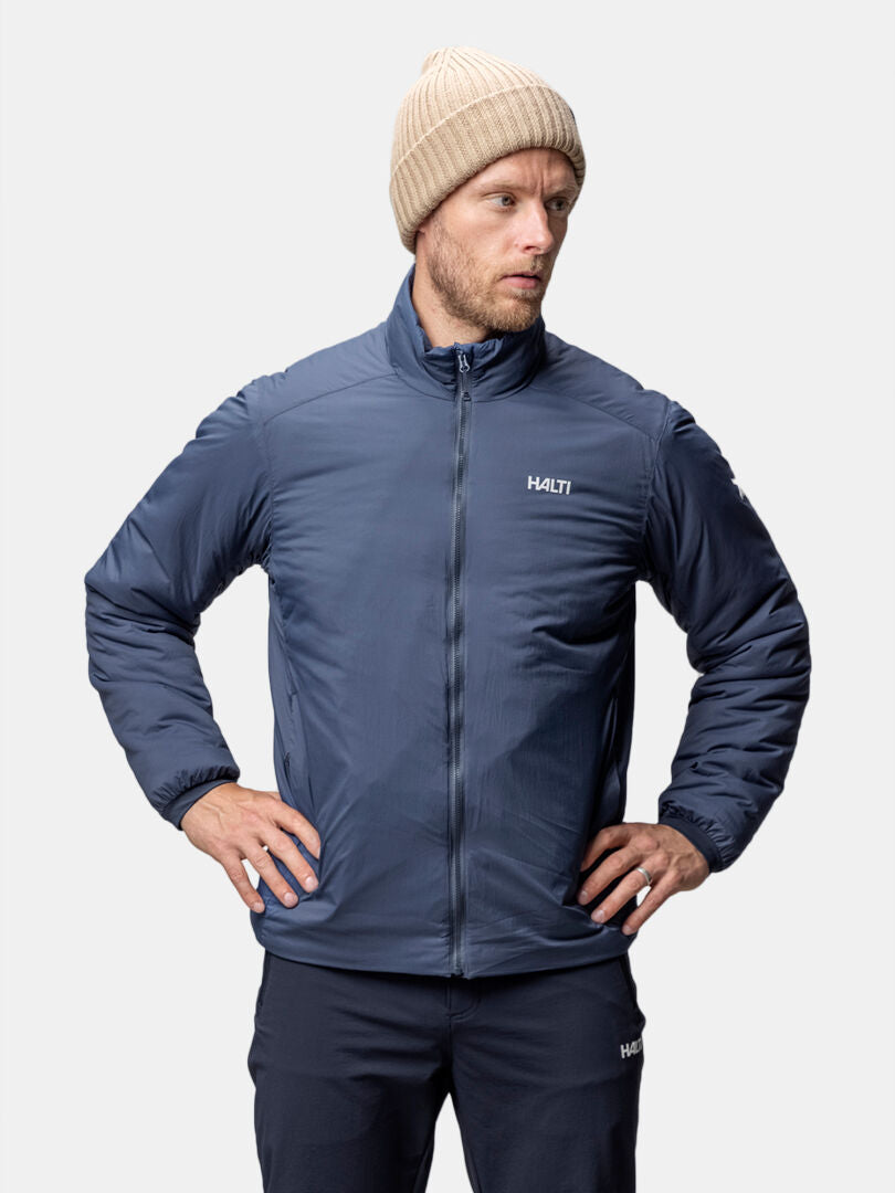 Mies, jolla on beige neuletakki, tummat housut ja sininen Halti Haave Primaloft Takki Miesten seisoo kädet lanteilla.
